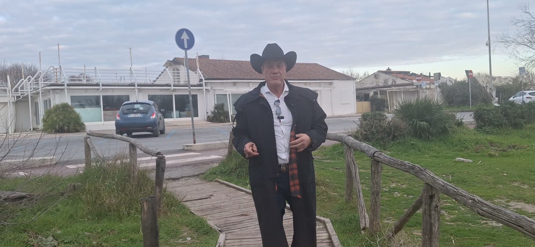 Tom Cummins walks a rustic boardwalk toward the camera in a cowboy hat and duster coat.