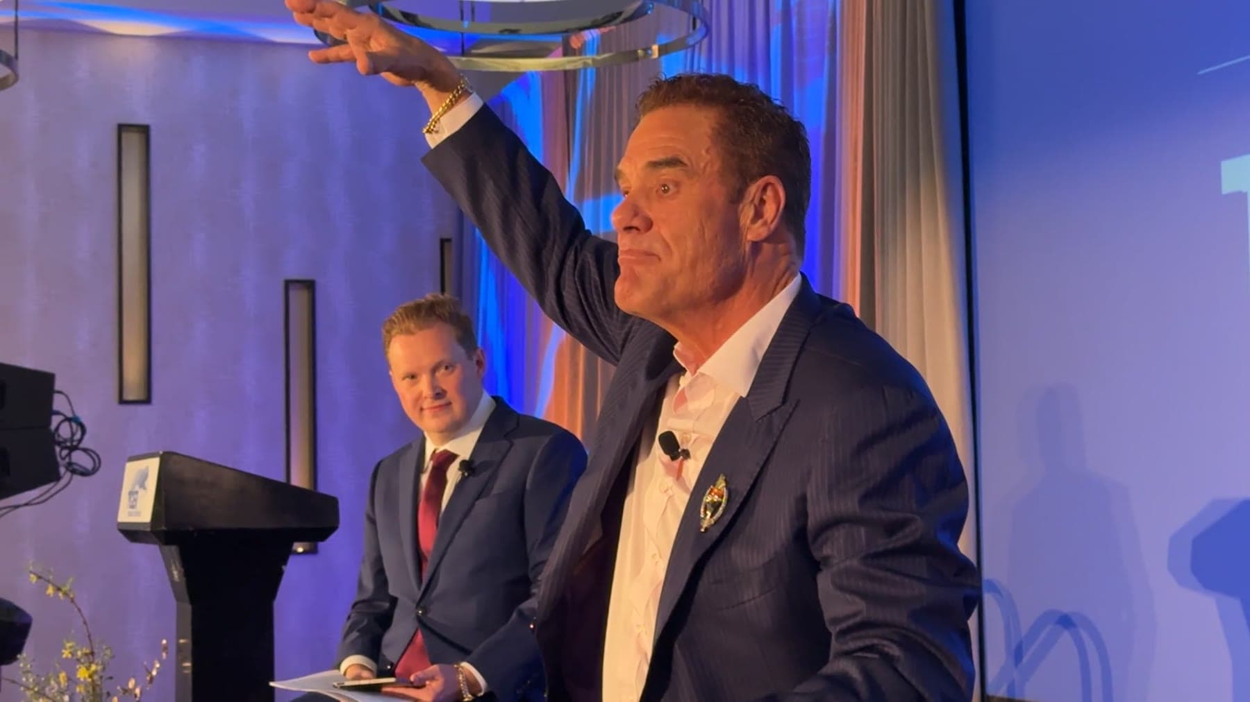 Tom Cummins sweeps an arm overhead while speaking on a stage lit in blue and purple.
