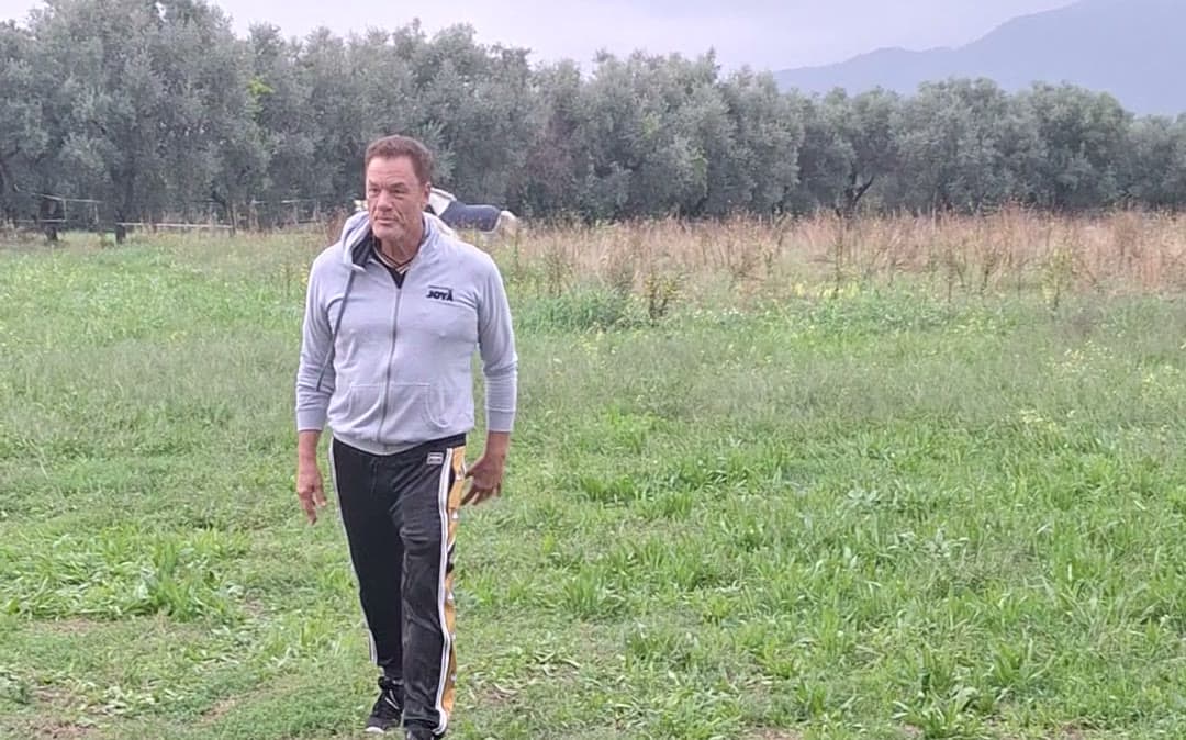 Tom Cummins walks across a pasture toward the camera with a horse grazing behind him in Tuscany.