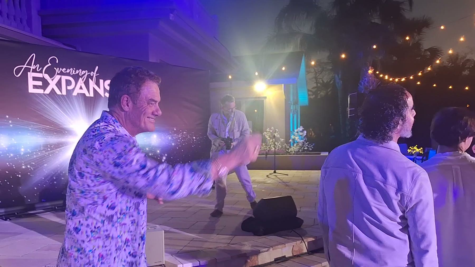 Tom Cummins grins mid-dance near the stage at an evening event while a band plays.
