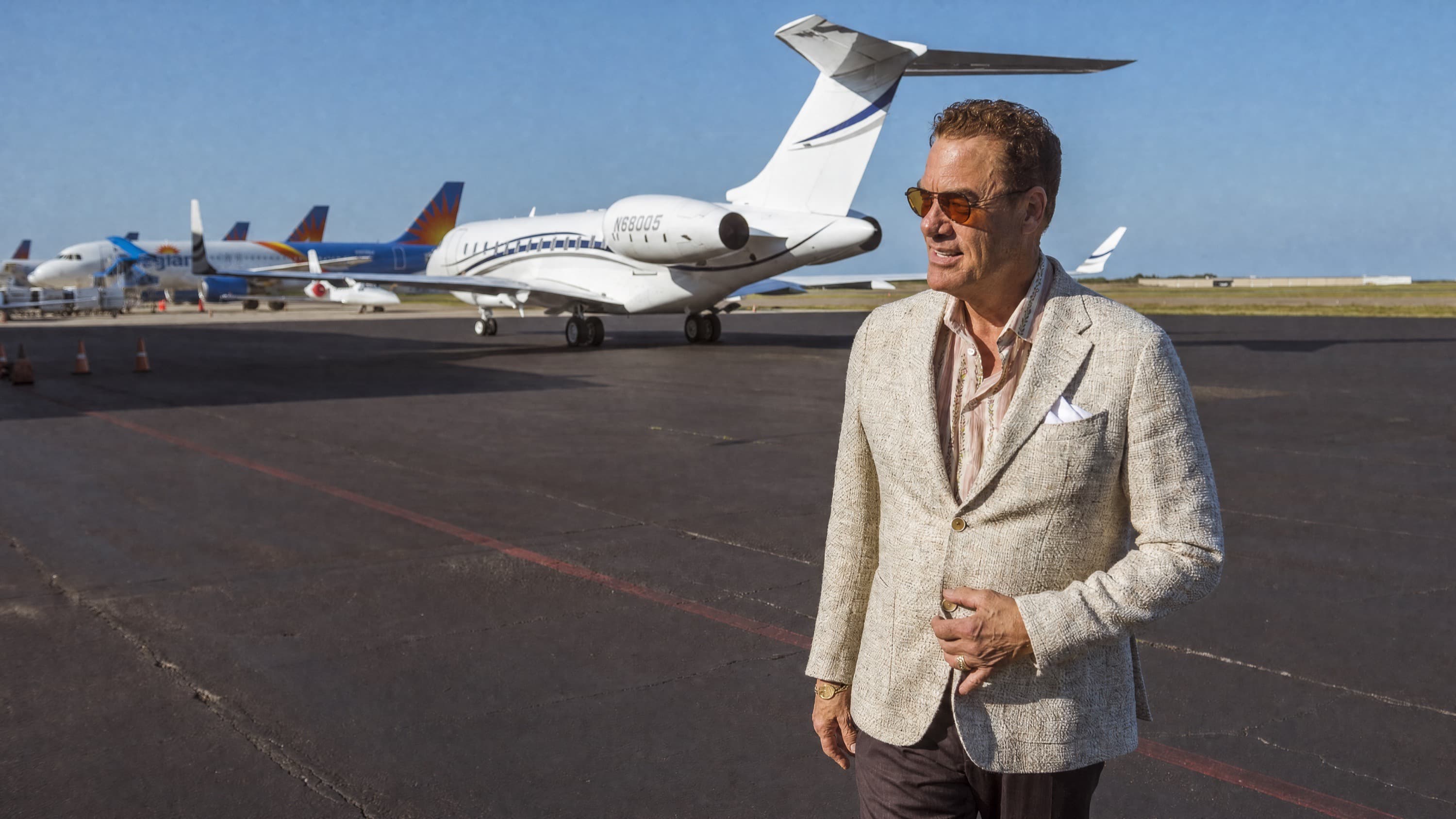 Tom Cummins walking the tarmac at golden hour in a cream tweed blazer, his private jet and a row of airliners behind him