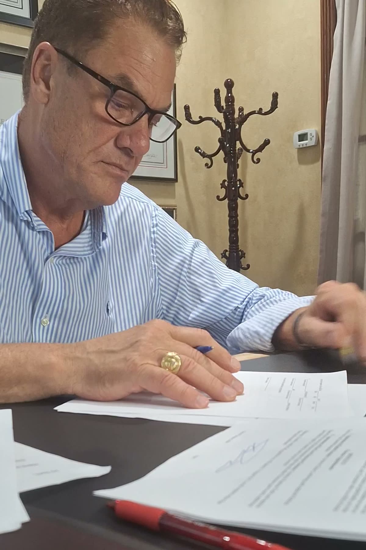 Tom in reading glasses working through and signing a stack of documents at his desk, red pen in the foreground