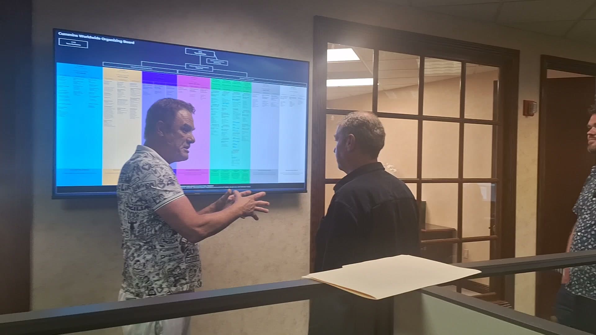 Tom mid-gesture briefing staff at a wall-size screen showing the company organizing board