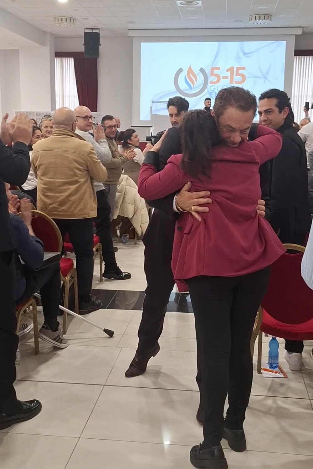 Tom lifting an attendee in a joyful embrace in the middle of a packed seminar hall in Bologna