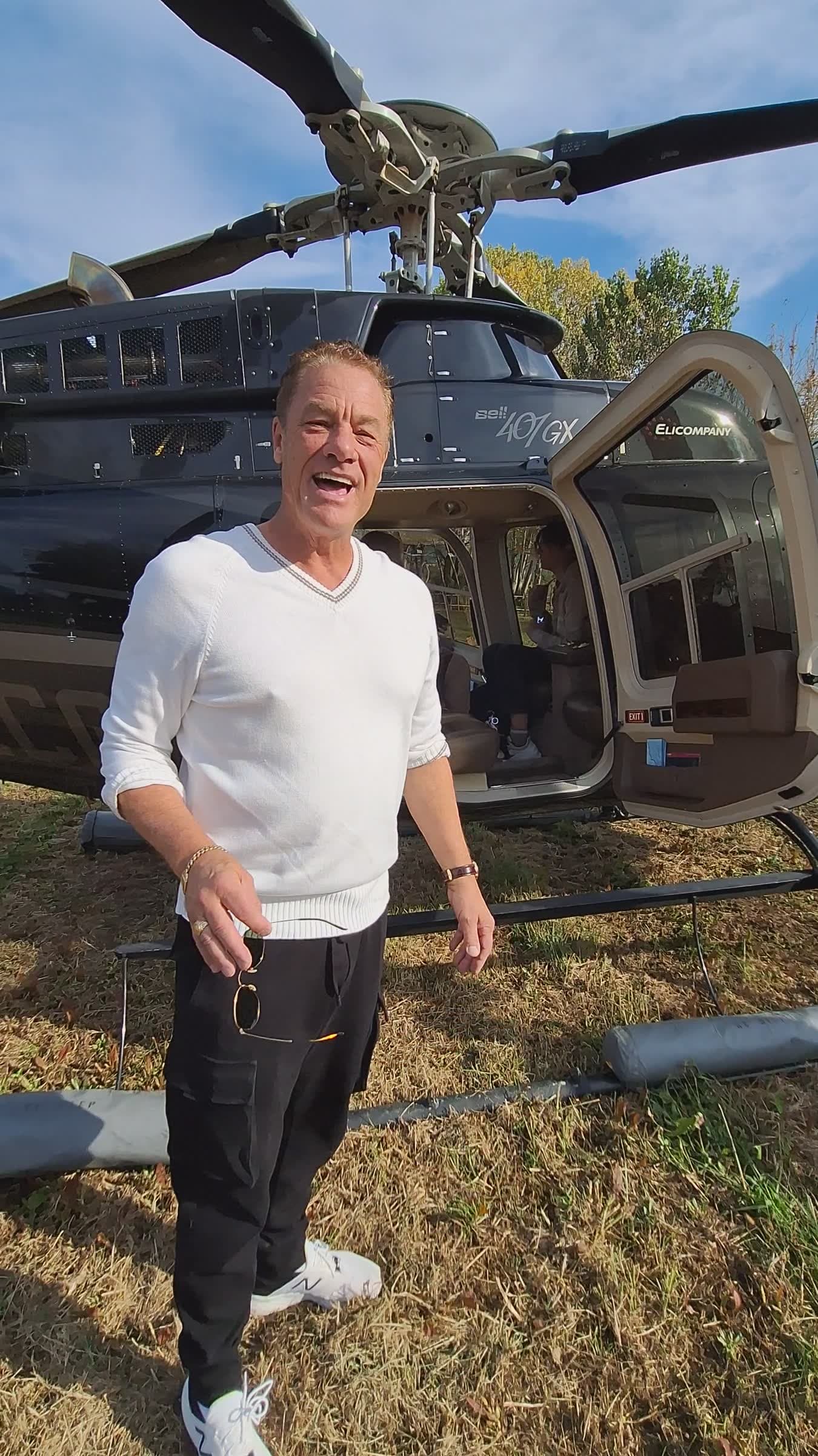 Tom Cummins beaming straight into the camera in a white sweater, a black Bell 407 helicopter with its cabin door open behind him in Tuscany