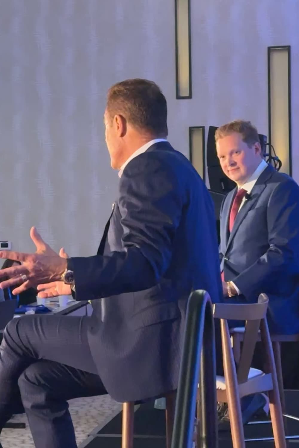 Tom Cummins seated on a stage stool mid-story, gesturing to the audience beside a moderator