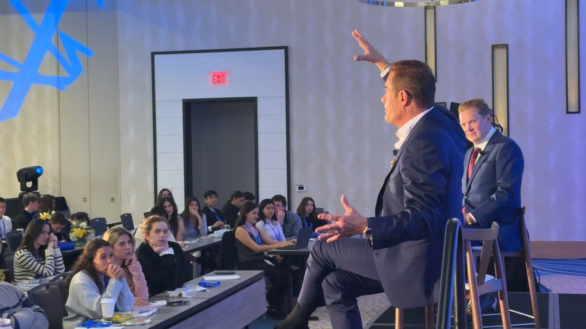 Tom Cummins addressing a full room from the stage with his arm outstretched.