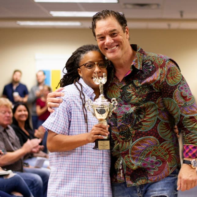 Tom with his arm around a team member holding a gold trophy as the office applauds