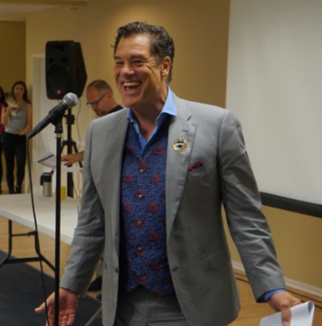 Tom laughing at the microphone in a gray blazer before a talk