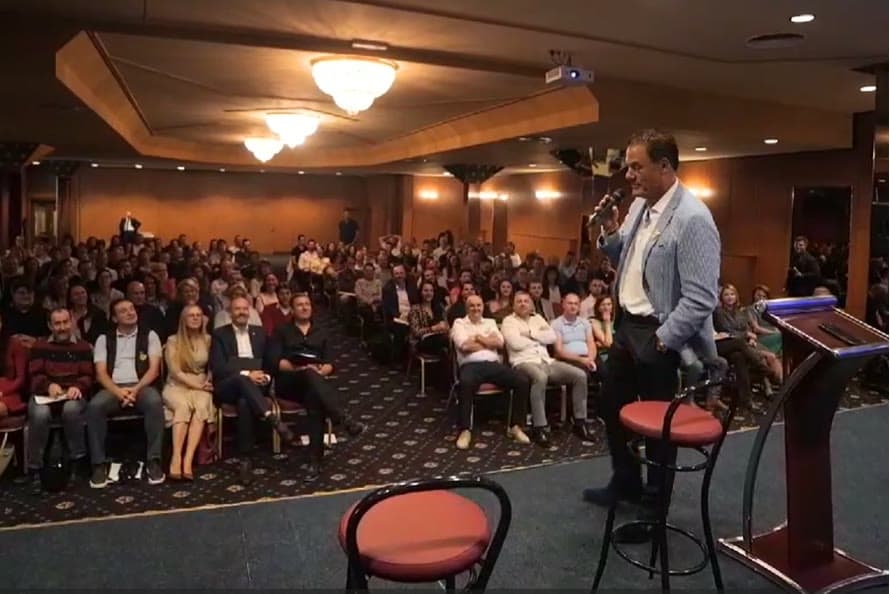 Tom on stage with a microphone addressing a full ballroom audience