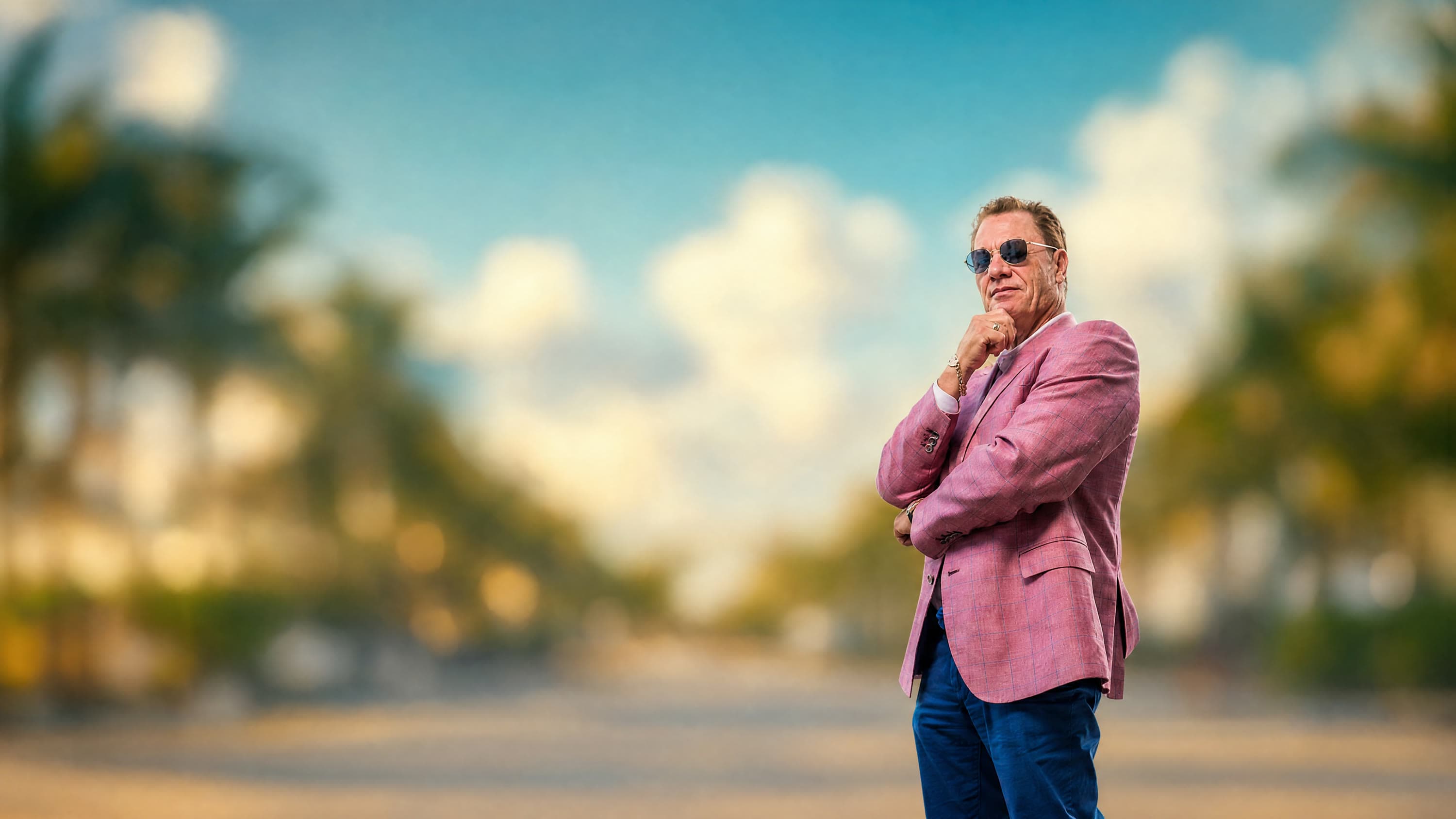 Tom Cummins in a pink blazer and sunglasses, hand on chin, on a sunlit boulevard